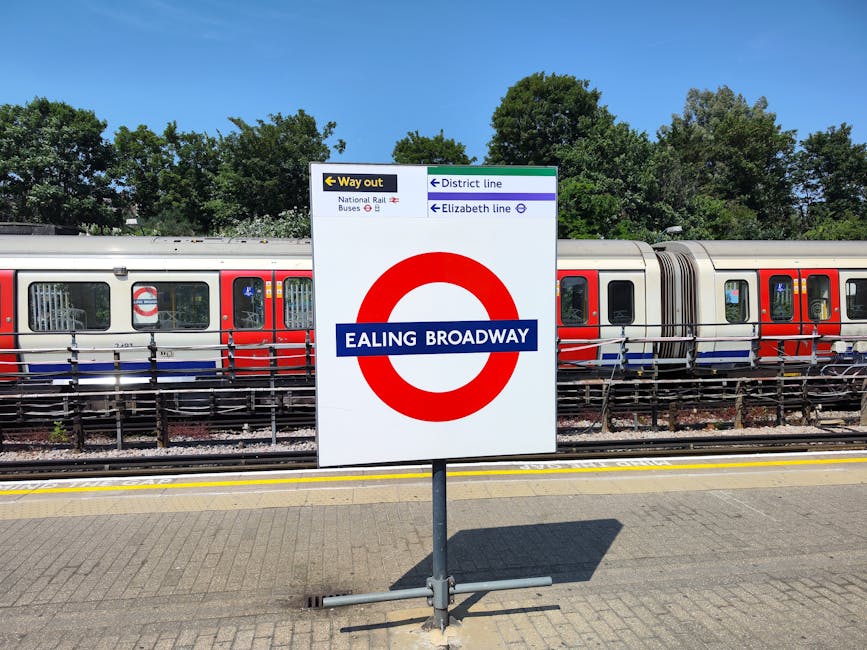 A sign for Ealing Broadway tube station displaying the station name in bold white letters on a blue background within a red circle, situated on a paved platform. Behind the sign, a modern London Under