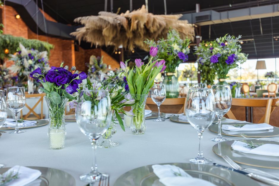 A table set for a floral event at the Colindale RAF Museum, featuring fresh flower arrangements in various glass vases with purple, lavender, and green blooms, including tulips and irises, arranged in