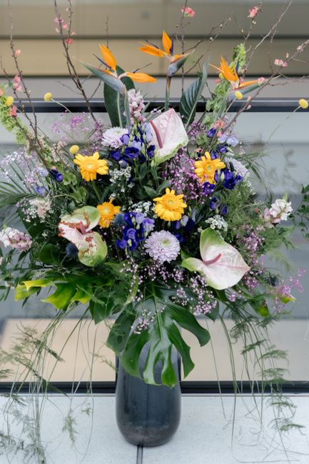 A lush floral arrangement featuring a variety of fresh flowers in a tall, dark vase. The bouquet includes vibrant orange Bird of Paradise flowers, white calla lilies with pink accents, purple and blue