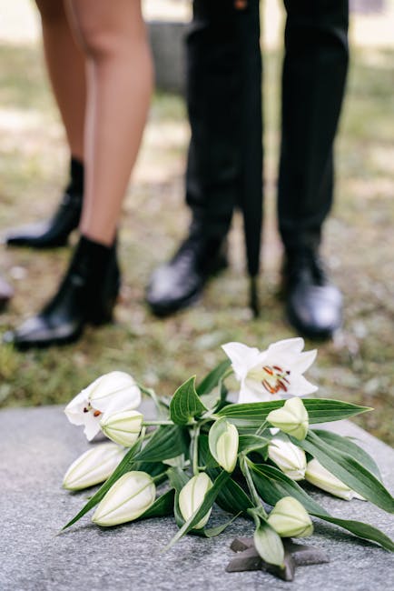 A delicate bouquet of white lilies and closed lily buds with long green leaves rests on a grey stone surface, featuring fresh, smooth petals and sturdy stems. In the background, the lower legs and fee