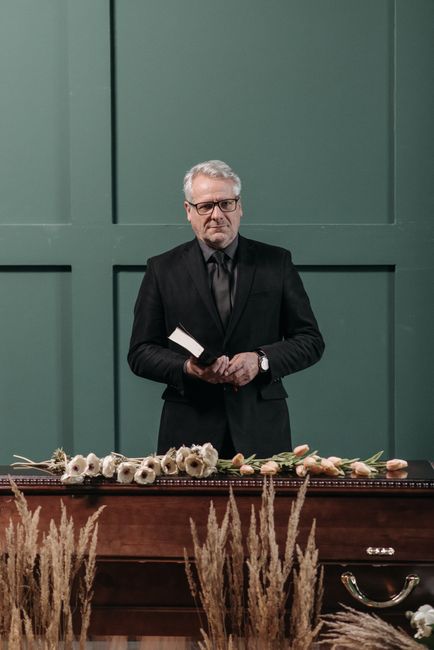 A man dressed in a black suit and glasses stands behind a dark wooden table, holding a white booklet in his left hand and looking contemplative. In front of him, a floral arrangement featuring white a