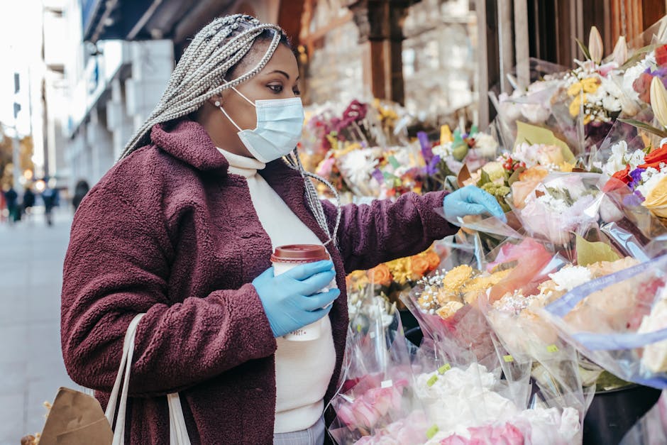 A woman wearing a face mask, blue gloves, and a burgundy coat is selecting a flower bouquet from an outdoor floral stall. The bouquet is wrapped in clear cellophane with pastel-colored paper, featurin
