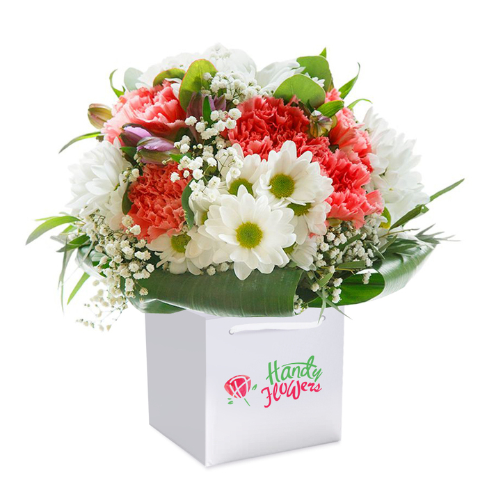 A meticulously composed arrangement-vivid in colour, lush in form-is displayed upright in a pristine square white gift bag, reflecting the sophisticated style of floristry found in Colindale. The bouquet's heart comprises coral and peach carnations, their generously ruffled petals radiating a soft, cheerful glow. Interspersed within are striking white daisies, each with a lime-yellow center that feels at once fresh and uplifting, like the atmosphere in Colindale's neighbourhood cafés. Dainty white sprigs of baby's breath dance between the blooms, lending an ethereal softness and textural variety. The entire bouquet is wrapped naturally by deep, shining green leaves, some broad and glossy, others more delicate, forming a verdant, protective rim. Look to the left, and you'll notice a few unopened buds tinged with purple-a gentle promise of blooms yet to open, reminiscent of the floral surprises spotted on walks along Colindale Avenue. The sturdy white ribbon handles and clean lines of the bag suggest ease of gifting and thoughtful presentation, perfect for a birthday, anniversary, or spontaneous delivery. Set against a seamless white background, every flower, leaf, and petal is accentuated by soft, even light, underscoring the freshness and artistry of local Colindale florists. This professional composition speaks to the care, attention, and celebratory spirit that define exceptional bouquet delivery within the area.